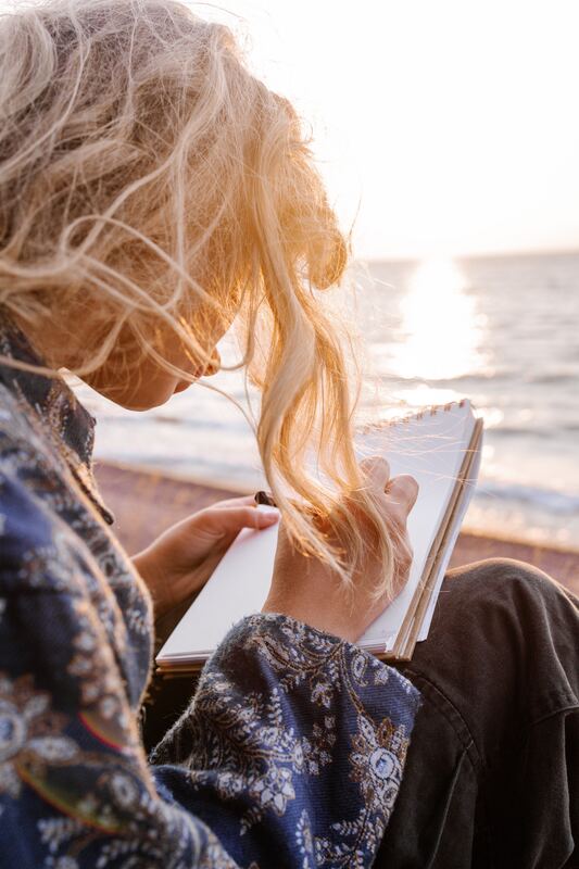 Girl at ocean sitting as the sun is rising.  She’s writing in her notebook, her blond hair pulled back, but long strands have become windblown. 