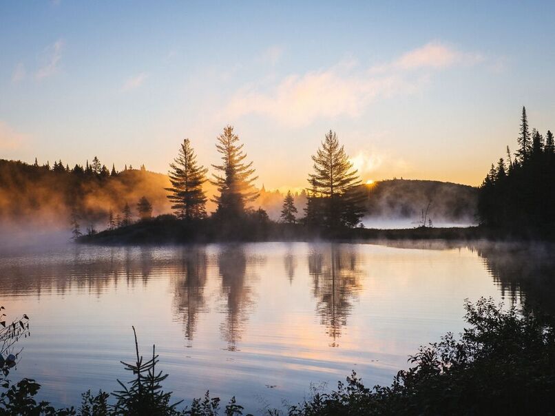 Winter landscape at sunrise over a lake with pines and mountains. Fog rising.