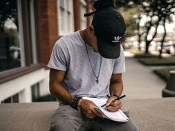 Young boy sitting on a sidewalk bench where a baseball cap, head down so we can’t see his face. He’s writing in a notebook. 